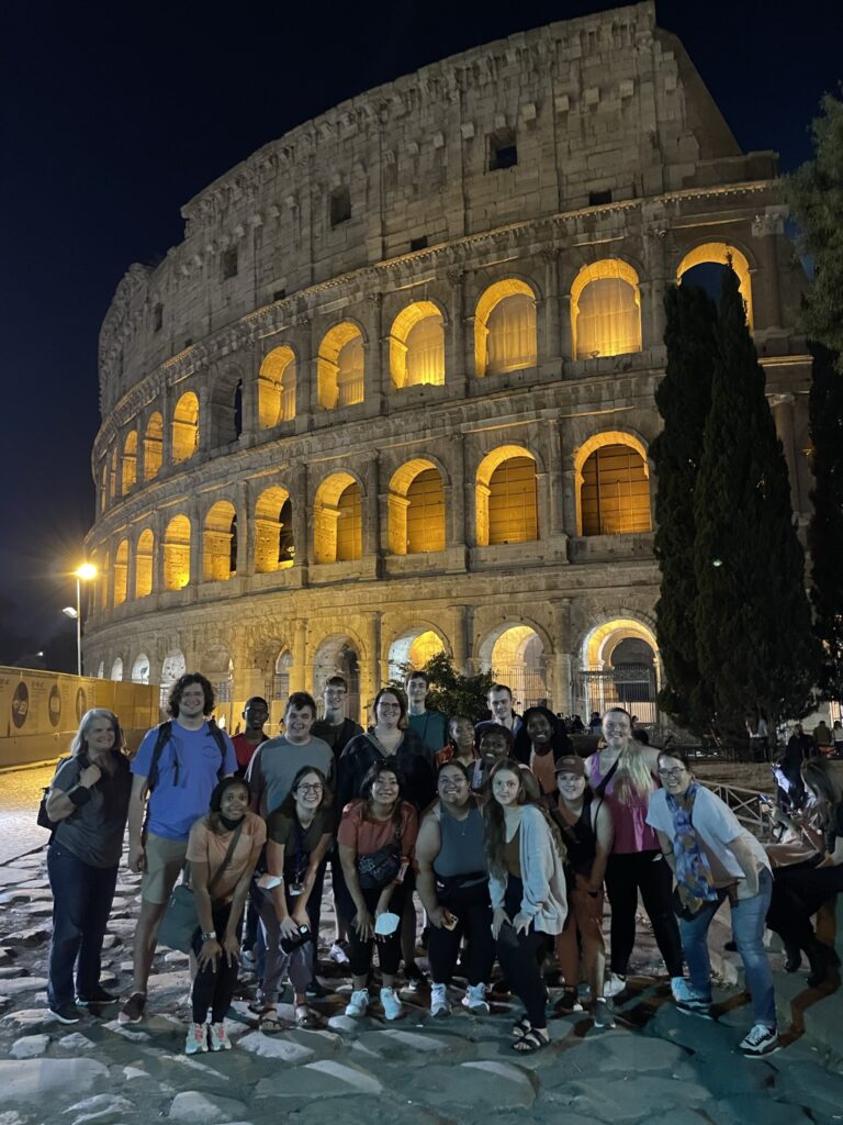 A group of students and instructors from the Classical Studies department at UNCG gathered in front of the Colosseum at night.