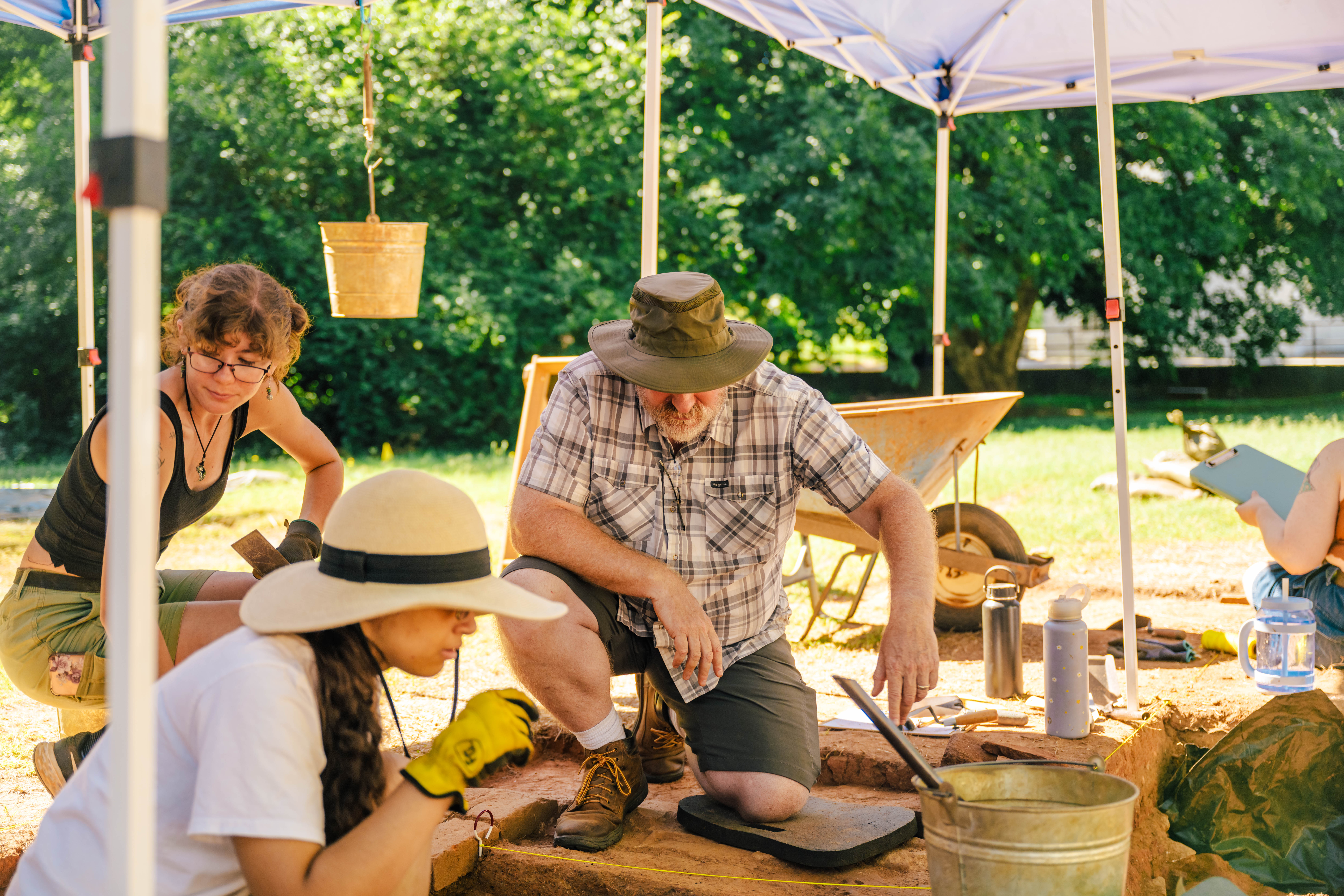 Man kneels to inspect an archaeological dig site covered with a tent as other student looks on.