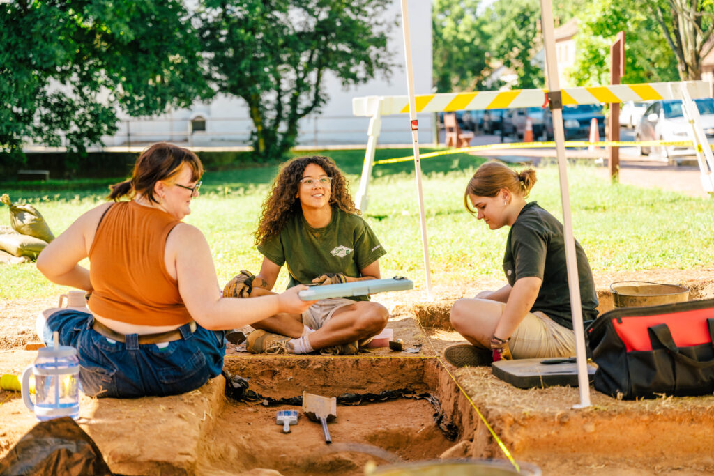 Three students sit on the ground with a measured archaeological dig site between them and tools like shovels and brushes all around.